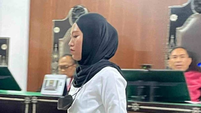 Terdakwa perempuan berhijab hitam dan kemeja putih sedang berdiri di ruang sidang Pengadilan Negeri Mataram.