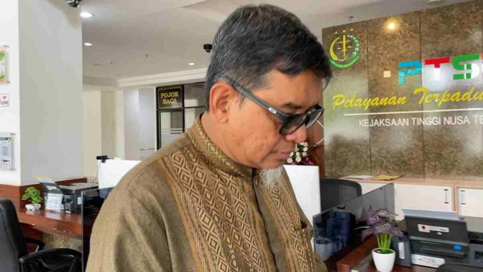 Logopit_1770968305314 Seorang pria paruh baya mengenakan kemeja batik cokelat dan kacamata hitam berdiri menunduk di depan meja pelayanan PTSP Kejaksaan Tinggi Nusa Tenggara Barat. Latar belakang menunjukkan interior kantor dengan logo Kejaksaan dan tulisan Pelayanan Terpadu Satu Pintu.