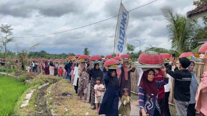 Iring-iringan warga perempuan di Lombok Timur berjalan di tepi sawah sambil menjunjung dulang dengan penutup anyaman berwarna merah di atas kepala.