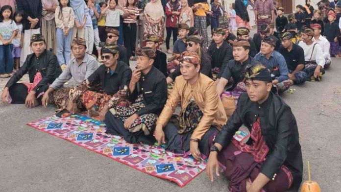 Sekelompok pria suku Sasak duduk bersila mengenakan pakaian adat hitam dan cokelat serta penutup kepala tradisional (sapuk) di sebuah jalan, dikelilingi oleh warga yang menyaksikan acara tersebut.