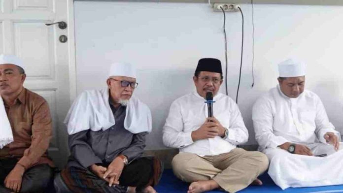 Logopit_1771288412801 Bupati Lombok Barat H. Lalu Ahmad Zaini mengenakan kemeja putih dan peci hitam sedang berbicara menggunakan mikrofon, duduk bersila di antara para tokoh agama yang mengenakan busana muslim dan peci putih.