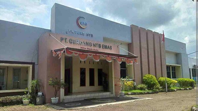 Foto tampak depan gedung kantor PT Gerbang NTB Emas dengan dominasi warna dinding krem dan abu-abu, terdapat papan nama perusahaan di bagian atas bangunan serta bendera Merah Putih di sisi kanan.