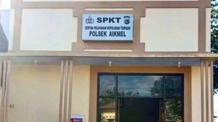 Papan nama SPKT Polsek Aikmel berwarna putih dengan logo kepolisian, terpasang di atas pintu kaca gedung kepolisian sebagai pusat layanan pengaduan masyarakat.