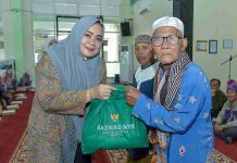 Wagub NTB Indah Dhamayanti Perkuat Jaring Pengaman Sosial di Sumbawa Wakil Gubernur NTB Indah Dhamayanti Putri mengenakan jilbab abu-abu memberikan tas bantuan hijau bertuliskan BAZNAS NTB kepada seorang lansia pria di dalam masjid.