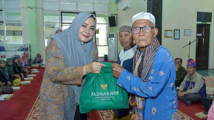 Wakil Gubernur NTB Indah Dhamayanti Putri mengenakan jilbab abu-abu memberikan tas bantuan hijau bertuliskan BAZNAS NTB kepada seorang lansia pria di dalam masjid.