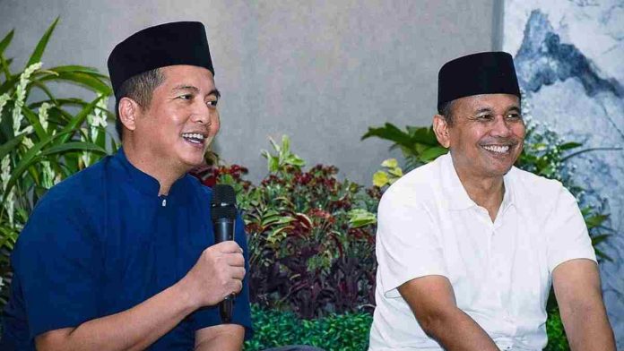 Gubernur NTB Lalu Muhamad Iqbal mengenakan baju koko biru dan peci hitam sedang memegang mikrofon, duduk berdampingan dengan pejabat pemprov dalam sebuah acara silaturahmi yang hangat.