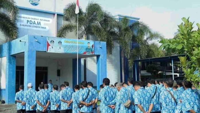 Barisan pegawai PDAM Kabupaten Lombok Timur mengenakan seragam batik biru sedang mengikuti apel pagi di halaman kantor yang bercat biru putih.