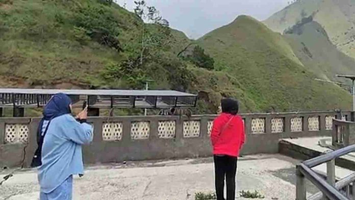 Dua orang wisatawan wanita, satu berjaket jeans biru dan satu berjaket merah, sedang berdiri di atas dek observasi beton sambil melihat pemandangan bukit-bukit hijau yang tinggi dan asri di Sembalun, Lombok Timur.