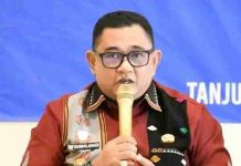 Lombok Utara Tekan Angka Stunting Lewat SOP Ketat Perkawinan Anak Foto close-up Kusmalahadi mengenakan kacamata, kemeja merah marun dengan selempang tenun Sasak Sasambo, memegang mikrofon emas di depan latar belakang biru bertuliskan "TANJUN..."
