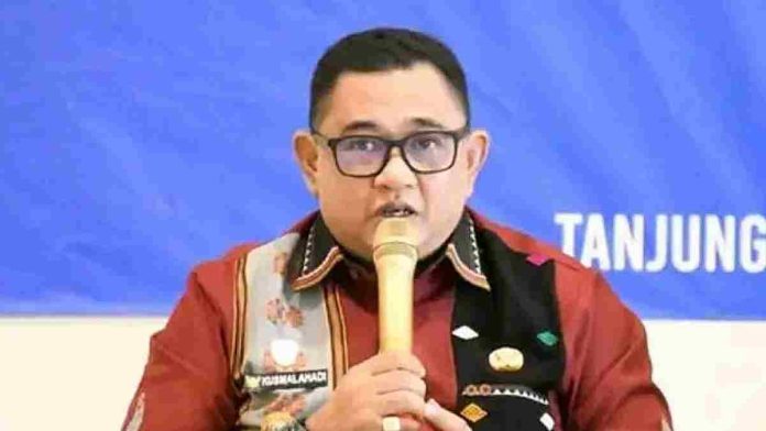 Foto close-up Kusmalahadi mengenakan kacamata, kemeja merah marun dengan selempang tenun Sasak Sasambo, memegang mikrofon emas di depan latar belakang biru bertuliskan 