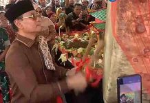 Mataram Heboh Aksi Spontan Wali Kota Mohan Roliskana Tabuh Beduk Bintaro Foto candid Wali Kota Mataram H. Mohan Roliskana mengenakan pakaian adat Sasak cokelat, kacamata hitam, dan peci hitam, sedang menabuh beduk besar di acara Lebaran Topat Makam Bintaro yang ramai pengunjung.