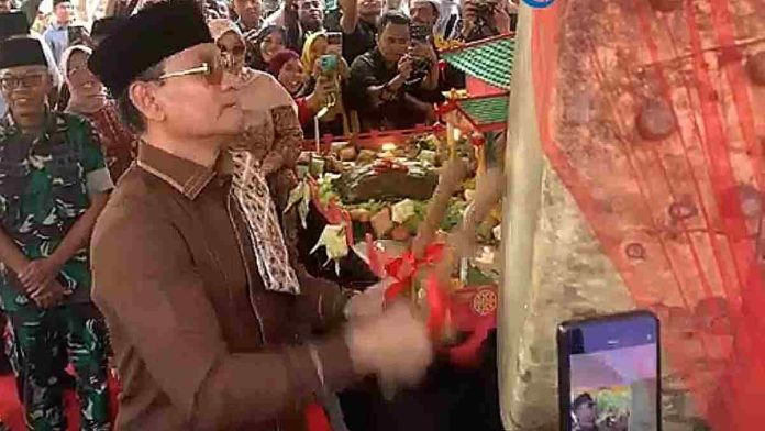 Foto candid Wali Kota Mataram H. Mohan Roliskana mengenakan pakaian adat Sasak cokelat, kacamata hitam, dan peci hitam, sedang menabuh beduk besar di acara Lebaran Topat Makam Bintaro yang ramai pengunjung.