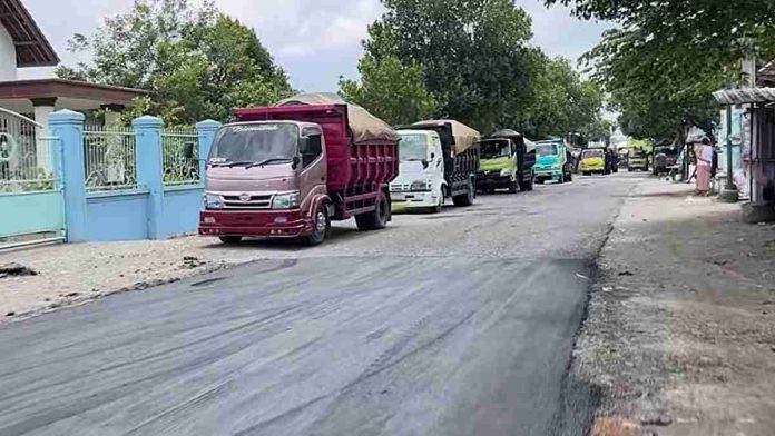 Truk-truk pengangkut material aspal terparkir berderet di pinggir jalan desa yang baru saja selesai diaspal mulus dengan latar belakang pepohonan dan rumah warga.