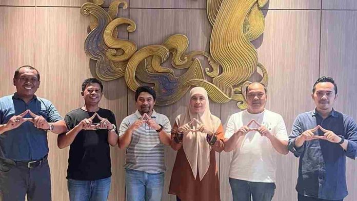 Enam orang pengurus DPC Partai Demokrat NTB berdiri berjejer sambil berpose membentuk simbol segitiga menggunakan jari tangan di depan latar dinding kayu dengan dekorasi ukiran emas.