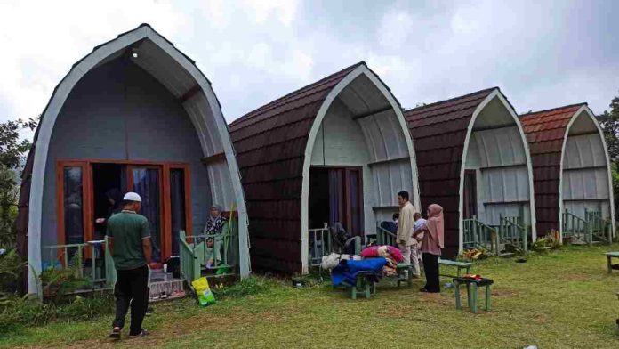 Deretan empat buah penginapan berbentuk kabin busur panah dengan atap genteng cokelat berjejer di area padang rumput hijau pegunungan Sembalun. Tampak beberapa orang wisatawan domestik sedang beraktivitas santai di depan teras dan halaman penginapan tersebut.