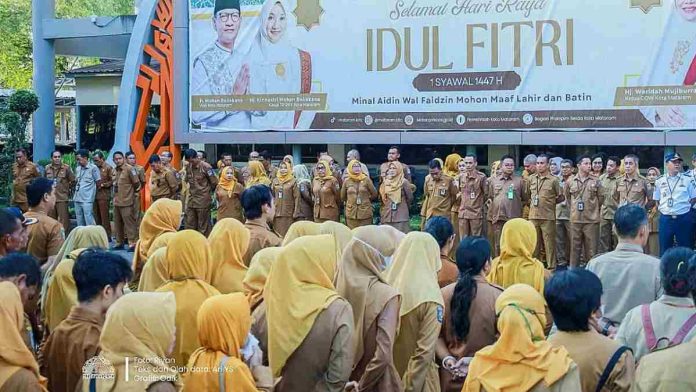 Kumpulan pegawai ASN Pemkot Mataram mengenakan seragam dinas cokelat (khaki) sedang berbaris rapi mendengarkan pidato apel pagi di lapangan dengan latar belakang baliho Idulfitri besar.