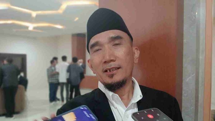 Pria dewasa mengenakan kopiah hitam, kemeja putih, dan jas hitam sedang berbicara di depan beberapa ponsel jurnalis dalam sebuah sesi wawancara di dalam ruangan.