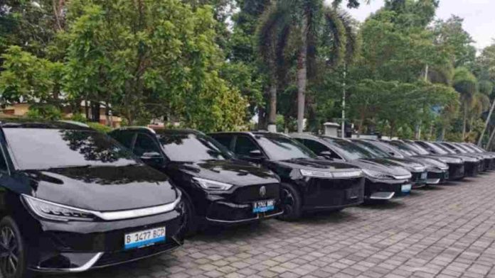 Barisan mobil listrik berwarna hitam terparkir rapi di area parkir terbuka dengan latar belakang pepohonan hijau di lingkungan kantor pemerintahan.