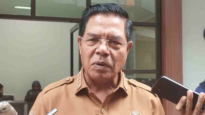 Foto close-up wajah Bupati Lombok Timur Haerul Warisin yang mengenakan seragam dinas cokelat dan kacamata, sedang memberikan keterangan kepada wartawan yang memegang smartphone.