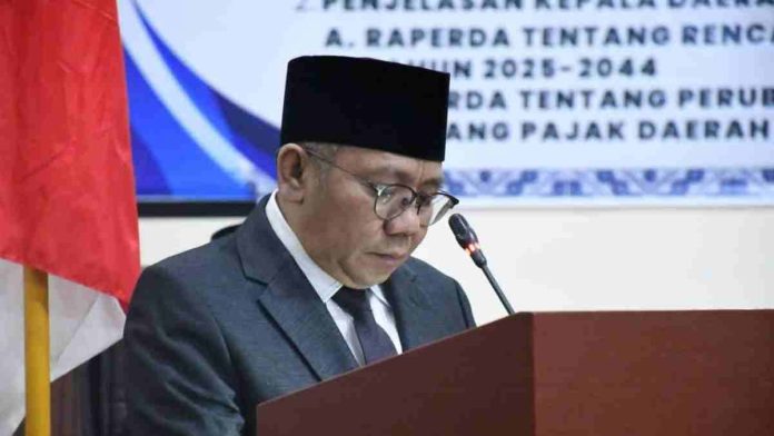 Bupati Lombok Utara Najmul Akhyar mengenakan jas abu-abu dan peci hitam, sedang membacakan dokumen LKPJ di podium dalam rapat paripurna DPRD.