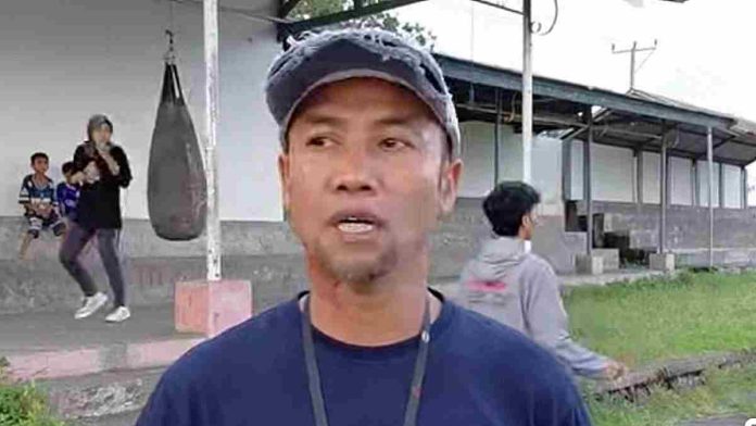 Foto close-up Lalu Gempar Satria mengenakan kaos biru gelap dan topi abu-abu lusuh, sedang berbicara dengan latar belakang area latihan bela diri di Pasar Bersih Masbagik.