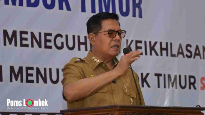Potret Bupati Lombok Timur, H. Haerul Warisin, berpidato mengenakan seragam dinas cokelat dan kacamata, berdiri di balik podium dengan latar belakang spanduk bertuliskan 