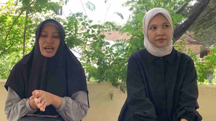 Dua wanita berhijab, Jamiatul Munawarah mengenakan kerudung hitam dan Baiq Restu mengenakan kerudung krem, sedang memberikan keterangan di luar ruangan dengan latar pepohonan hijau.