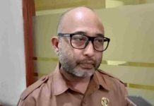 DLH Kota Mataram Tunda Kenaikan Tarif Retribusi Sampah Demi Warga Foto wajah H. Nizar Denny Cahyadi, Kepala DLH Kota Mataram, mengenakan seragam ASN cokelat dan kacamata hitam, sedang memberikan wawancara di depan pintu kaca.