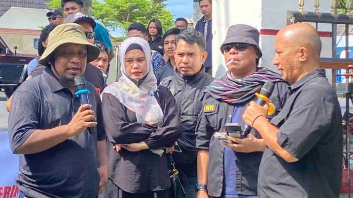 Sekelompok aktivis sedang melakukan orasi di depan umum; tampak seorang pria bertopi krem dan seorang pria berkepala plontos memegang mikrofon memberikan pernyataan di depan massa.