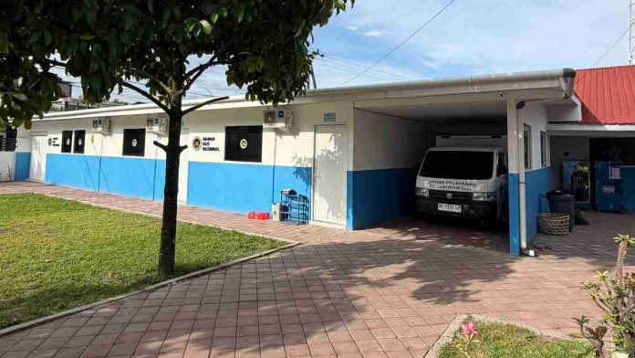 Pemandangan luar gedung kantor Satuan Pelayanan Program Gizi dengan cat dinding putih biru, halaman berpaving, dan sebuah mobil operasional terparkir di garasi terbuka.