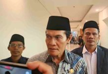 Pemkab Lombok Tengah Ajak Karang Taruna Keroyok Masalah Pernikahan Anak Foto narasumber Muhammad Nursiah mengenakan kemeja batik biru dan peci hitam sedang memberikan keterangan kepada media di koridor hotel dengan latar belakang beberapa staf.