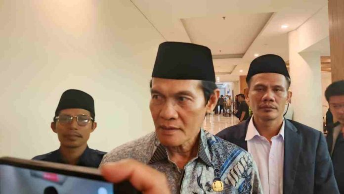 Foto narasumber Muhammad Nursiah mengenakan kemeja batik biru dan peci hitam sedang memberikan keterangan kepada media di koridor hotel dengan latar belakang beberapa staf.