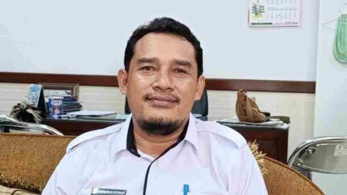 Foto narasumber Sosiawan Putraji, seorang pria mengenakan kemeja putih dengan papan nama di dada, duduk di ruangan kantor dengan latar belakang meja kerja dan kalender dinding.