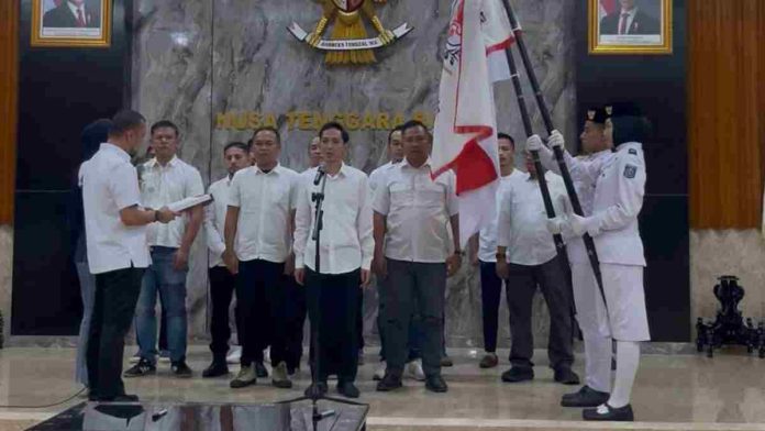 Sejumlah pria berpakaian kemeja putih berdiri tegak di dalam ruangan formal saat prosesi pelantikan, didampingi petugas pembawa bendera organisasi di sisi kanan.