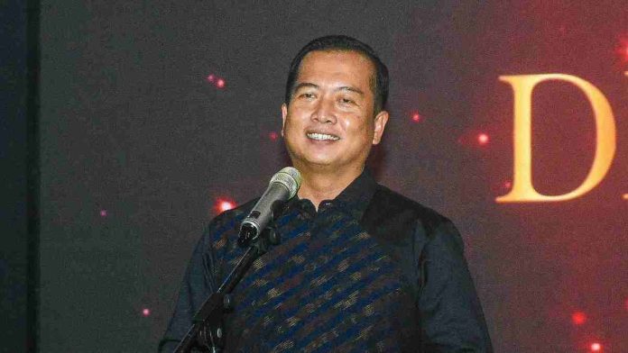 Foto close-up Gubernur NTB H. Lalu Muhamad Iqbal mengenakan kemeja batik bermotif garis diagonal, sedang tersenyum dan berbicara di depan mikrofon dalam acara formal malam hari.