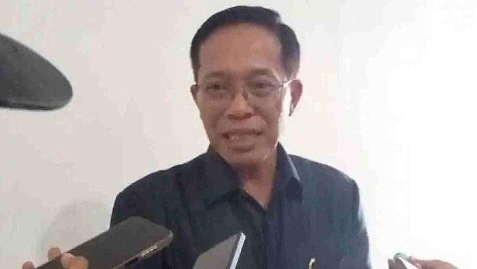 Foto close-up Sekretaris Daerah Lombok Timur, M. Juaini Taofik, berkacamata dan mengenakan kemeja gelap, sedang berbicara di depan sejumlah mikrofon dan ponsel wartawan.