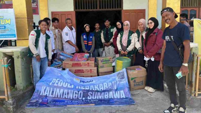 Sekelompok relawan mengenakan jaket hijau dan rompi biru berfoto bersama warga di depan tumpukan bantuan logistik berupa kardus mi instan, kompor gas, dan ember di Desa Kalimango.