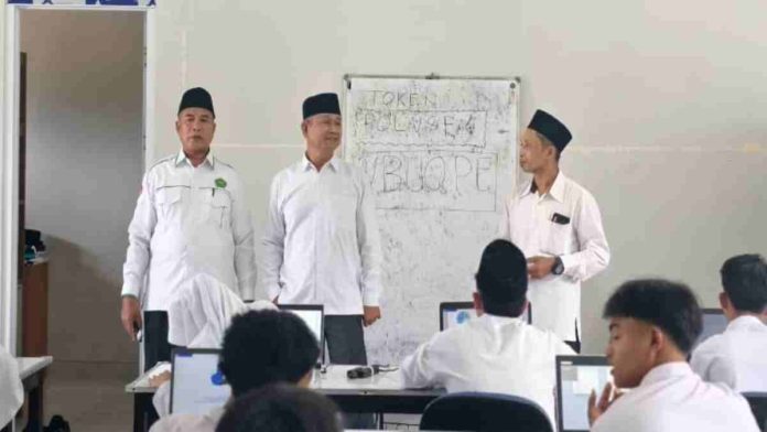 Tiga orang pria mengenakan kemeja putih dan peci hitam berdiri di depan kelas memantau siswa yang sedang mengerjakan ujian di depan komputer masing-masing. Di belakang mereka terdapat papan tulis putih.