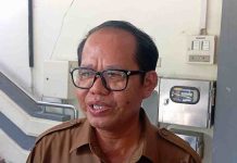 Nomenklatur Nama SD dan SMP se-Kota Mataram Resmi Berubah Tahun Ini Foto close-up Kepala Dinas Pendidikan Kota Mataram, M. Yusuf, mengenakan seragam cokelat (ASN) dan kacamata hitam, sedang berbicara dengan latar belakang tangga gedung.
