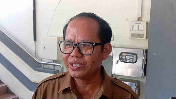 Foto close-up Kepala Dinas Pendidikan Kota Mataram, M. Yusuf, mengenakan seragam cokelat (ASN) dan kacamata hitam, sedang berbicara dengan latar belakang tangga gedung.