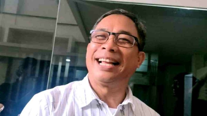 Foto close-up Muhammad Aminurlah (Haji Maman), pria berkacamata dengan kemeja putih motif garis tipis, sedang tertawa lebar dengan latar belakang pantulan kaca gedung.