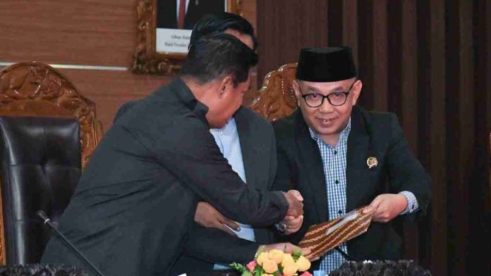Bupati Lombok Timur H. Haerul Warisin mengenakan jas hitam dan peci, sedang bersalaman sambil menerima map cokelat dari pimpinan DPRD di atas meja sidang yang dihiasi bunga.