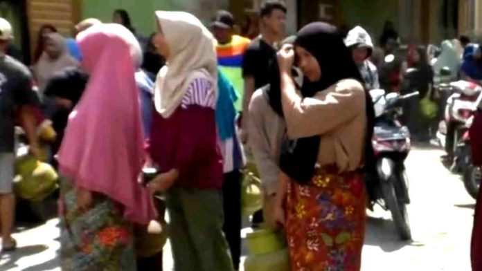 Sekelompok mahasiswa berjaket almamater hijau dan warga berdiri di depan tumpukan bantuan logistik seperti kardus mie instan, kompor gas, dan baskom plastik di bawah spanduk 