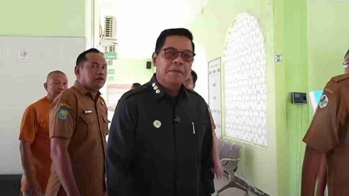 Bupati Lombok Timur H. Haerul Warisin mengenakan seragam dinas hitam dan kacamata, berjalan di dalam koridor puskesmas atau rumah sakit diikuti oleh beberapa staf berseragam ASN cokelat.