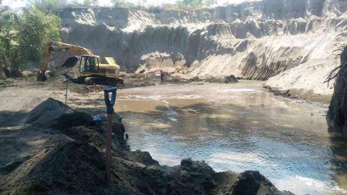 Aktivitas tambang galian C di area tebing pasir yang luas, terlihat satu unit ekskavator kuning sedang beroperasi di dekat genangan air cokelat.