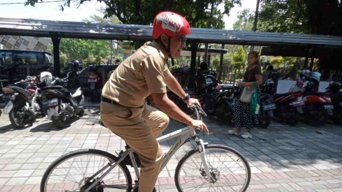 Seorang pria berseragam ASN cokelat mengenakan helm merah sedang mengendarai sepeda perak di area parkir yang dipenuhi deretan sepeda motor.