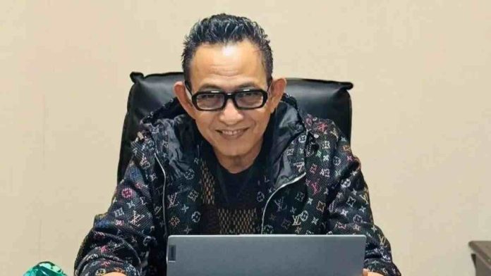 Foto potret Andi Syarifuddin, kuasa hukum terdakwa, mengenakan jaket bermotif dan kacamata hitam, duduk di depan laptop dengan ekspresi tersenyum ke arah kamera.