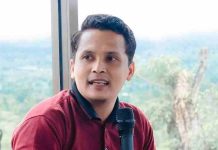 Ade Putra Tepis Isu Ulat Menu Makan Bergizi di Lombok Timur Foto Ade Putra Kurniawan mengenakan kaos merah marun sedang berbicara memegang mikrofon dengan latar belakang jendela besar yang memperlihatkan pemandangan perbukitan.