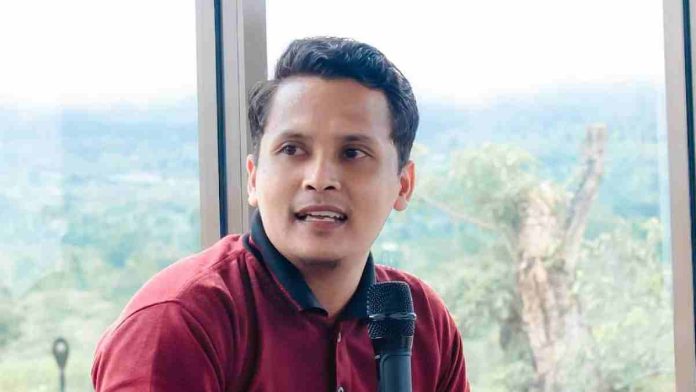 Foto Ade Putra Kurniawan mengenakan kaos merah marun sedang berbicara memegang mikrofon dengan latar belakang jendela besar yang memperlihatkan pemandangan perbukitan.