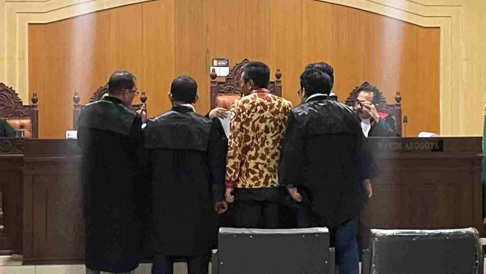 Suasana persidangan tipikor di mana terdakwa berbaju batik berdiri di hadapan majelis hakim bersama para penasihat hukumnya untuk memeriksa dokumen.